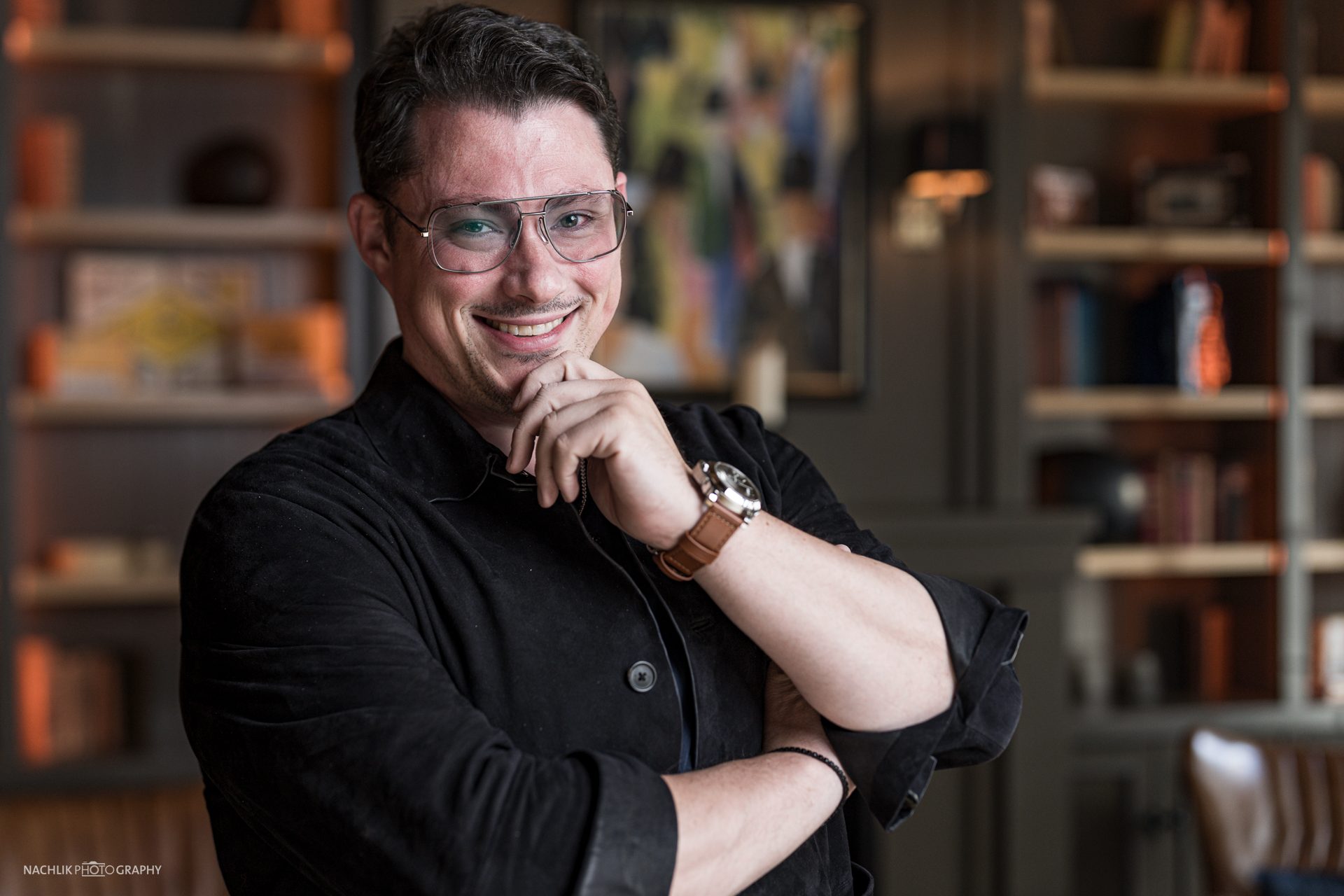 Ricardo Marinello lächelnd vor einer Bücherwand, Hand am Kinn
